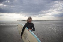Mujer mayor caminando desde el mar, llevando tabla de surf - foto de stock