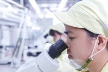 Mujer joven usando microscopio en la estación de control de calidad en la fábrica produciendo placas de circuitos electrónicos flexibles. Planta se encuentra en el sur de China, en Zhuhai, provincia de Guangdong - foto de stock