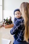 Woman carrying baby son wearing knit hat in kitchen — Stock Photo