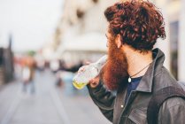 Молодой хипстер с рыжими волосами и бородой пьет пиво в бутылках на городской улице. — стоковое фото
