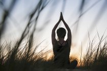 Vue arrière de la femme adulte moyenne pratiquant le yoga dans de longues herbes silhouettées au coucher du soleil — Photo de stock