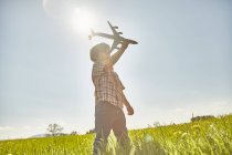 Boy in sunlit field with blue sky playing with toy airplane — Stock Photo
