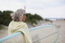 Donna anziana che cammina verso la spiaggia, portando la tavola da surf, vista posteriore — Foto stock
