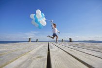 Young girl running on wooden pier, holding bunch of balloons — Stock Photo