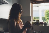 Молода жінка має перерву на каву, дивлячись через кухонне вікно — стокове фото