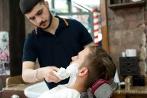 Junger Mann im Friseursalon trägt Rasierschaum auf das Gesicht der Kunden auf — Stockfoto