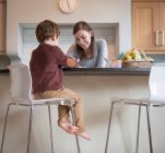 Boy sitting on stool with mother in kitchen and drawing — Stock Photo