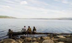 Три туриста сидят в ряд на упавшем дереве у озера — стоковое фото