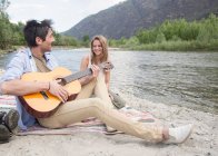 Друзья сидят у реки, играют на гитаре — стоковое фото