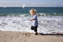Niño corriendo desde el agua en la orilla del mar con yates en la distancia - foto de stock