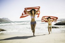 Mother and daughter are running on beach holding towel over head — Stock Photo