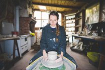 Vista frontal de la joven en el taller sentada a la rueda de cerámica haciendo olla de barro, mirando hacia abajo - foto de stock