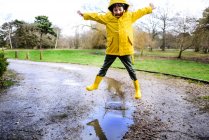 Boy in yellow anorak jumping above puddle in park — Stock Photo