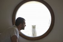 Середній дорослий чоловік дивиться на кота через кругле вікно — стокове фото