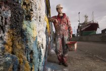 Pintor de navio masculino segurando tinta pode encostar contra a parede salpicada de tinta — Fotografia de Stock