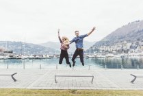 Parejas jóvenes en paseo marítimo saltando en el aire, Lago de Como, Italia - foto de stock