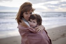 Mother and daughter wrapped in blanket hugging on beach — Stock Photo