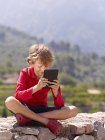 Niño sentado en la pared de piedra mirando tableta digital, Mallorca, España - foto de stock