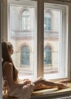 Mid adult woman sitting on window ledge, looking out of window — Stock Photo