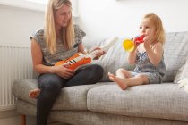 Female toddler and mother playing with toy trumpet and guitar on sofa — Stock Photo