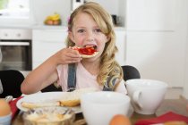 Chica comiendo tostadas con mermelada en la mesa de la cocina y mirando en la cámara - foto de stock
