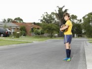 Портрет угрюмого мальчика, держащего футбольный мяч на пригородной дороге — стоковое фото