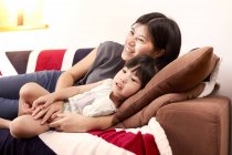 Young Chinese mother and daughter laying on sofa watching the television together at home — Stock Photo