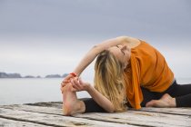 Metà donna adulta toccando dita dei piedi praticare yoga sul molo di legno del mare — Foto stock