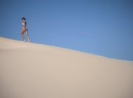 Jeune femme marchant sur une dune de sable contre un ciel bleu clair — Photo de stock