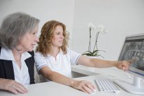 Зрелая женщина показывает пожилой женщине рентгеновское изображение, указывающее на экран компьютера — стоковое фото