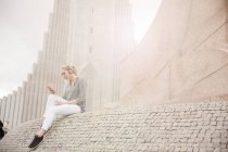 Blondhaarige junge erwachsene Frau sitzt in der Nähe des Kirchturms und hört Musik, reykjavik, Island — Stockfoto