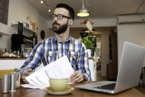 Homme client lecture de la paperasse et en utilisant un ordinateur portable à la table de café — Photo de stock