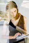 Mujer joven usando tableta digital, fotografiada a través de vidrio - foto de stock