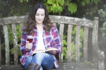 Mujer adulta leyendo y bebiendo vino tinto en un banco de jardín en el castillo de Thornbury, South Gloucestershire, Reino Unido - foto de stock