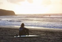 Joven surfista masculino sentado mirando desde la playa, Devon, Inglaterra, Reino Unido - foto de stock