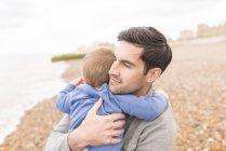 Отец обнимает маленького сына на галечном пляже — стоковое фото
