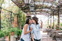 Lesbisches Paar in bepflanztem Torbogen macht Selfie mit Smartphone, küsst sich auf Wange, Florenz, Toskana, Italien — Stockfoto