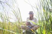 Середня доросла жінка сидить у довгій траві, використовуючи цифровий планшет — стокове фото