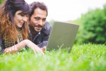 Cabeza y hombros de pareja joven acostada en la hierba usando computadora portátil sonriendo - foto de stock