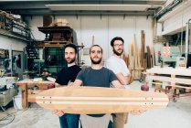 Friends standing in carpentry workshop holding skateboard looking at camera — Stock Photo