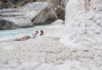 Young men relaxing on rocky beach using smartphone, Golfo di Orosei, Sardinia, Italy — Stock Photo