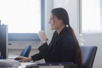 Vista laterale della giovane donna in ufficio seduta alla scrivania usando il computer sorridente — Foto stock