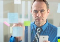 Businessman with smartphone sticking notes on office glass wall — Stock Photo
