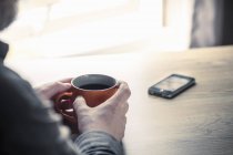 Immagine ritagliata di uomo che tiene una tazza di caffè a tavola con smartphone — Foto stock