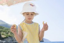 Menina usando chapéu de sol comer donut na praia — Fotografia de Stock