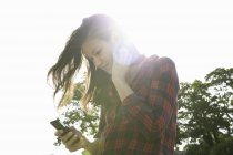 Jovem mulher lendo textos de smartphones contra o céu iluminado pelo sol — Fotografia de Stock