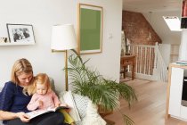 Woman and toddler daughter reading book on living room sofa — Stock Photo
