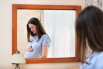 Pregnant woman holding stomach, standing in front of mirror — Stock Photo