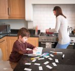 Niño para colorear en el libro en el mostrador de la cocina como madre prepara la cena - foto de stock