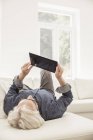 Старший лежит на диване, с помощью цифрового планшета, вид сзади — стоковое фото